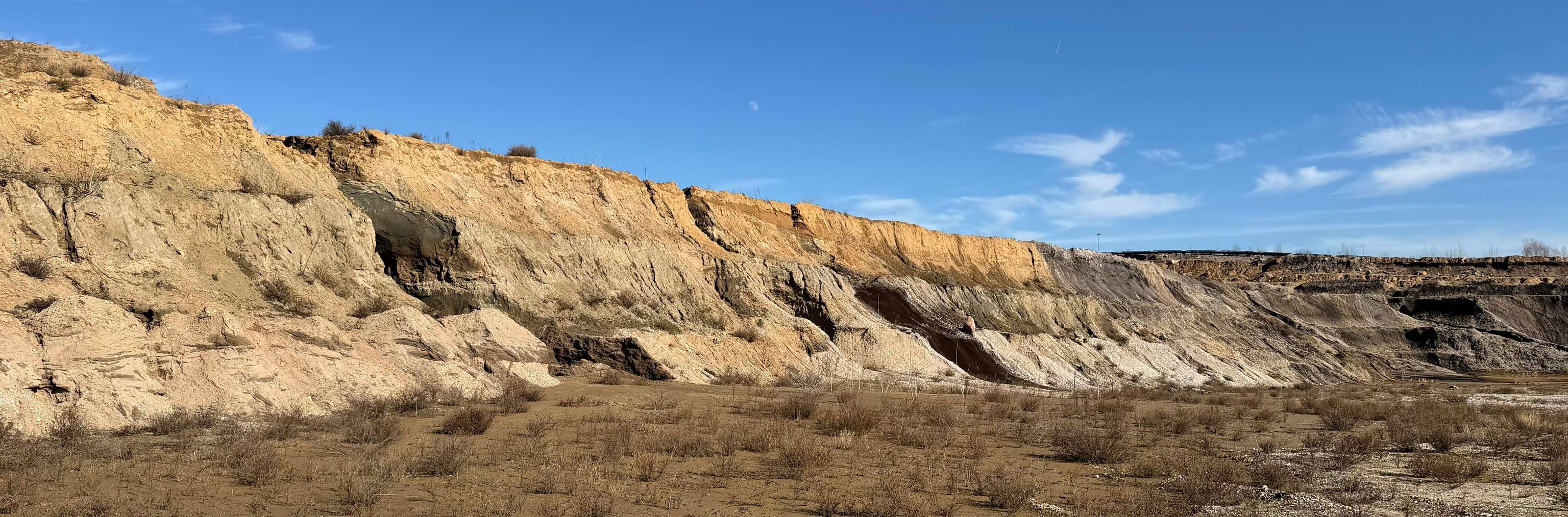 Erosionslandschaft mit steilen, sandigen Felswänden und trockenem Boden unter blauem Himmel.