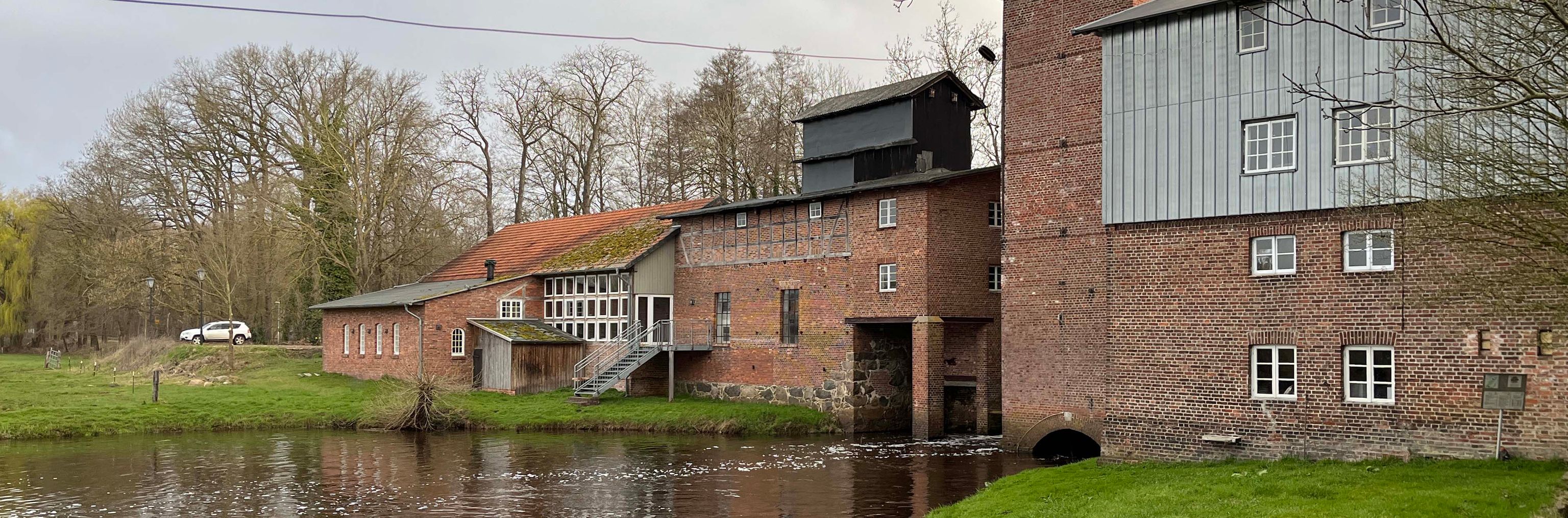 Altes Backsteingebäude am Wasser mit grünem Hintergrund und überdachtem Bereich.